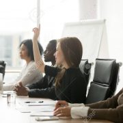 Smiling curious businesswoman raising hand at group meeting voti Smiling curious young businesswoman raising hand at multiracial group meeting engaging in offered activity, voting as volunteer or asking question at corporate business training, seminar or workshop